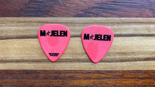 Two red guitar picks with black 'MAJELEN' and 'Shred Picks' branding, placed on a striped wood grain surface.