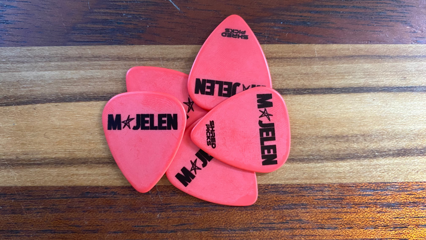 Five red guitar picks with black printed text and logos, including 'MAJELEN' and 'Shred Picks', arranged on a wooden surface.